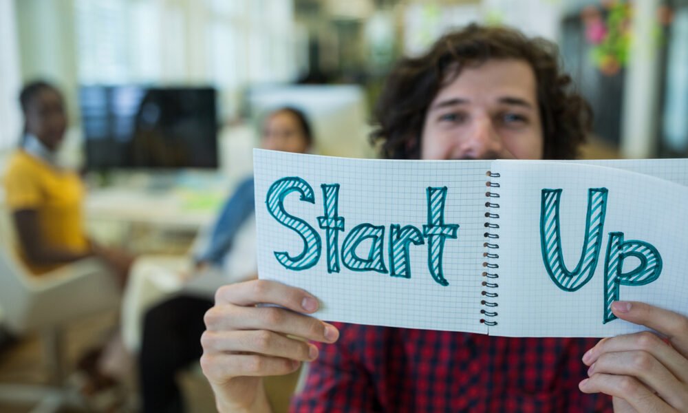 elinde “Start Up” yazılı not defteri tutan kişi ve arka planda ofis ortamı, girişimcilik ve start-up kültürünü temsil eden konsept görsel