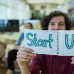 Bir Girişim Ne Zaman Başarılı Sayılır? Exit, Sürdürülebilirlik ve Etki elinde “Start Up” yazılı not defteri tutan kişi ve arka planda ofis ortamı, girişimcilik ve start-up kültürünü temsil eden konsept görsel