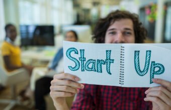 elinde “Start Up” yazılı not defteri tutan kişi ve arka planda ofis ortamı, girişimcilik ve start-up kültürünü temsil eden konsept görsel